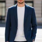 Muž v modrom saku a bielom tričku stojí na rozmazanom mestskom pozadí za denného svetla